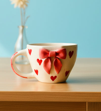Hand holding a decorative mug with red bows and hearts against a light blue background